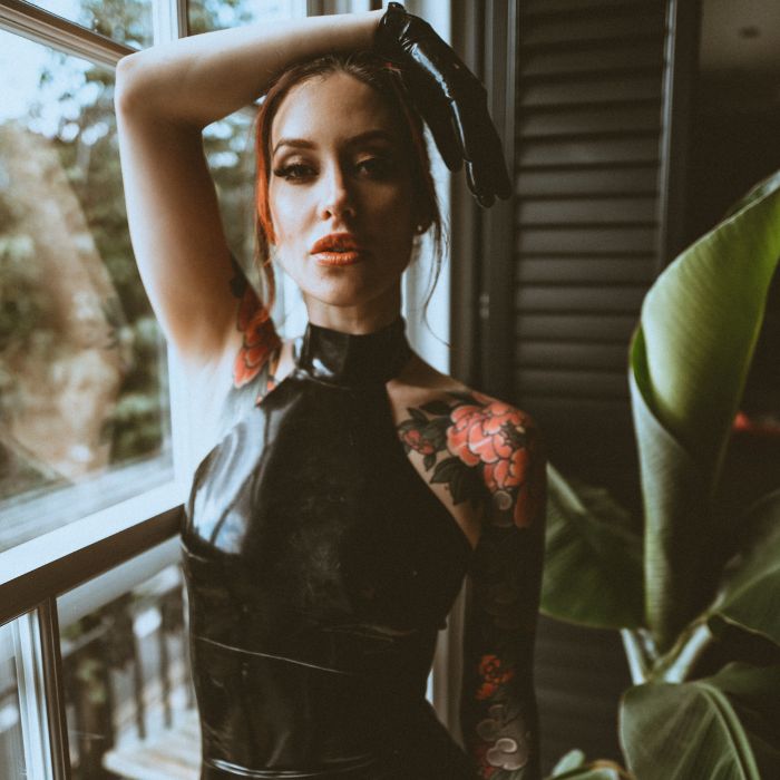 Mistress Adreena in a latex dress with one arm over her head.