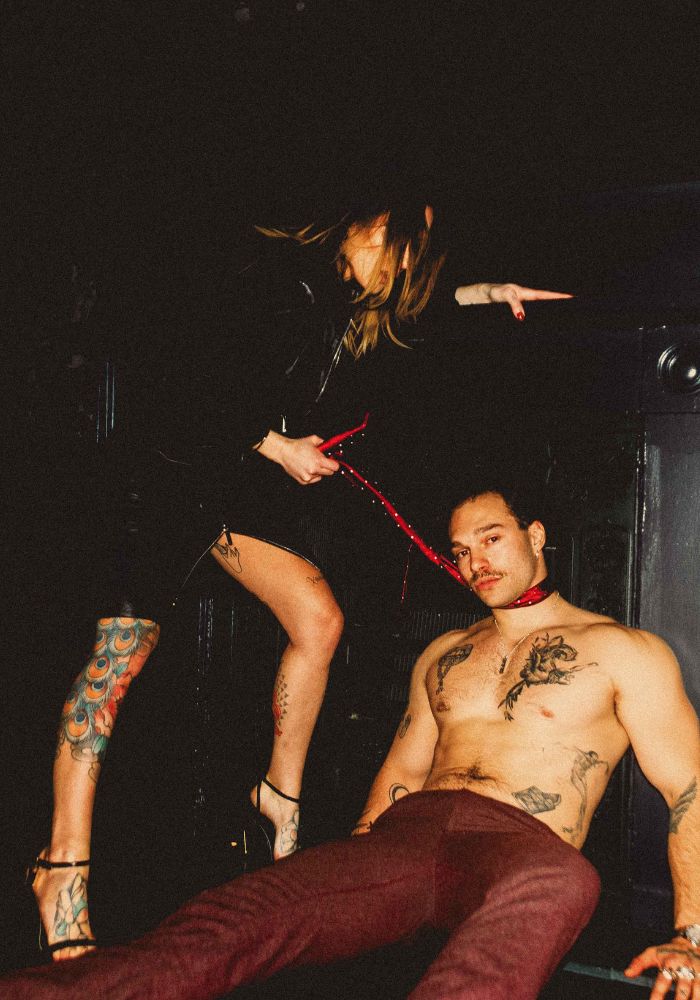 Mistress Adreena standing above her submissive, pulling his tie.