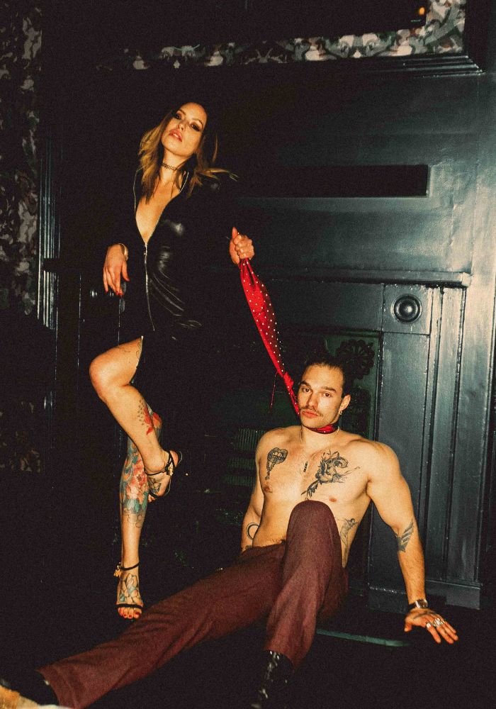 Mistress Adreena standing above her submissive, pulling his tie.