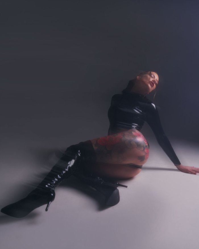 Woman with floral leg tattoos wearing black latex and high boots sitting on the floor against dark background.