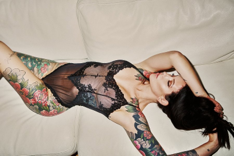 Woman with colorful tattoos lying on a white couch wearing a sheer black lace bodysuit.