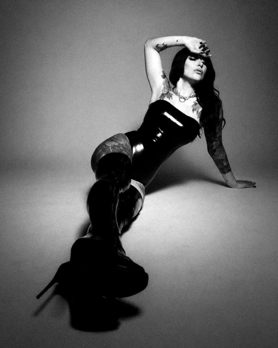 Black and white photo of a tattooed woman in latex dress and boots reclining on the floor, hand on forehead.