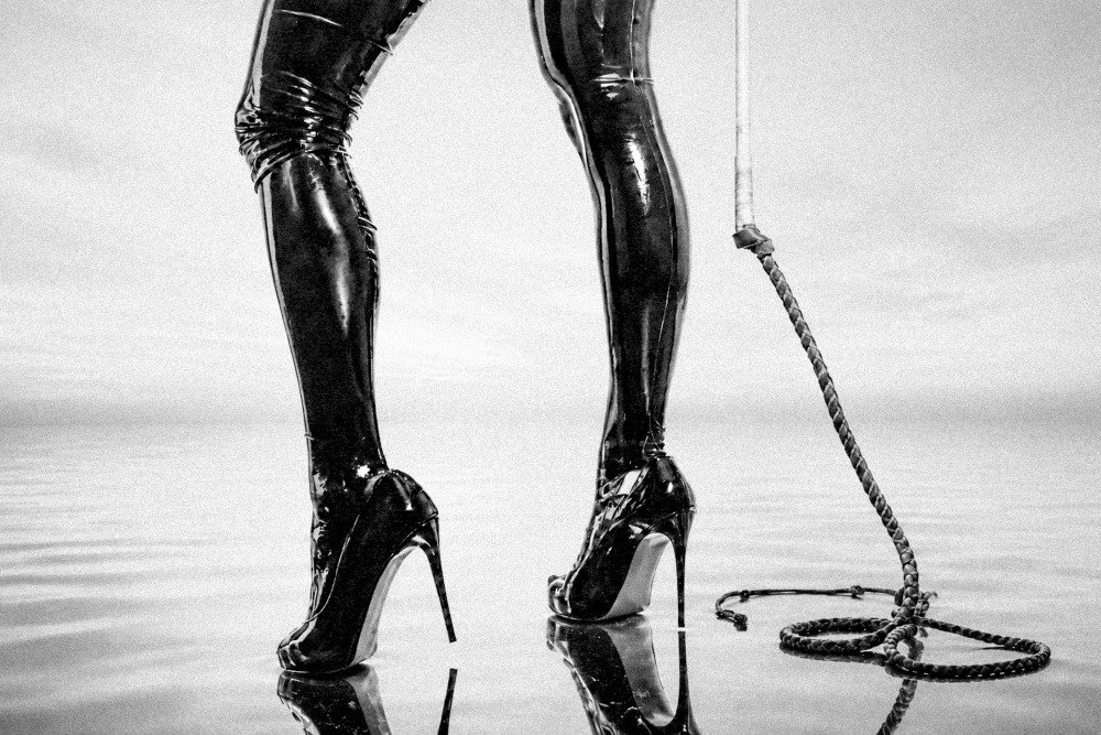 Black high-heeled boots with shiny leather pants standing on a reflective surface next to a coiled whip.