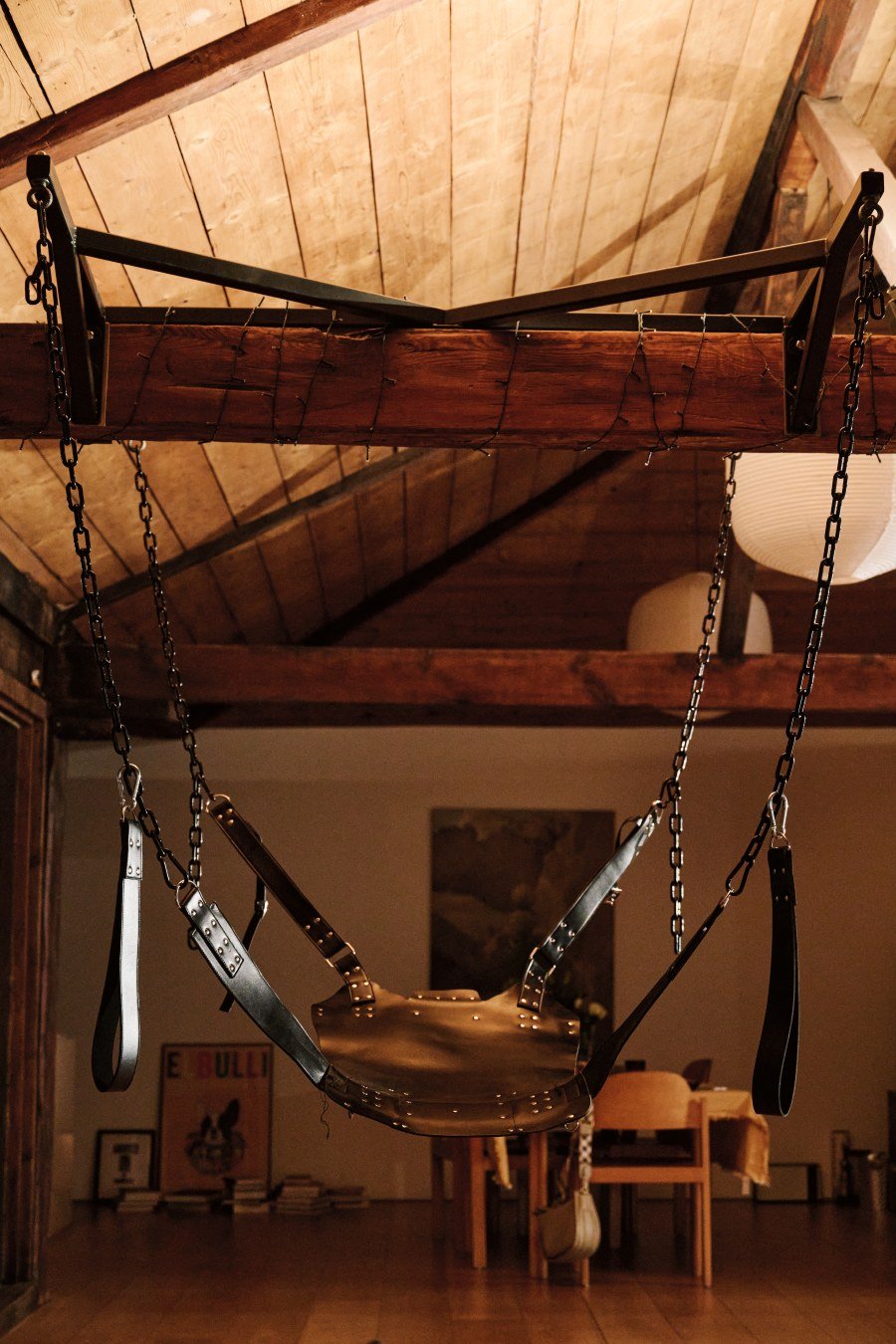 Black leather swing suspended by chains from a wooden beam in a warm-toned room.