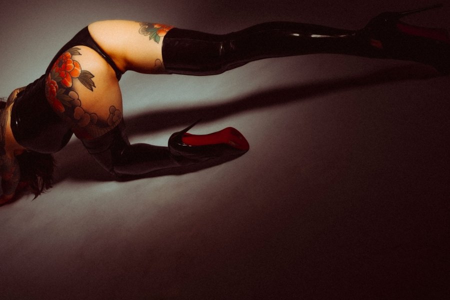 Person in black latex thigh-high boots and tattooed legs posing dramatically on the floor.