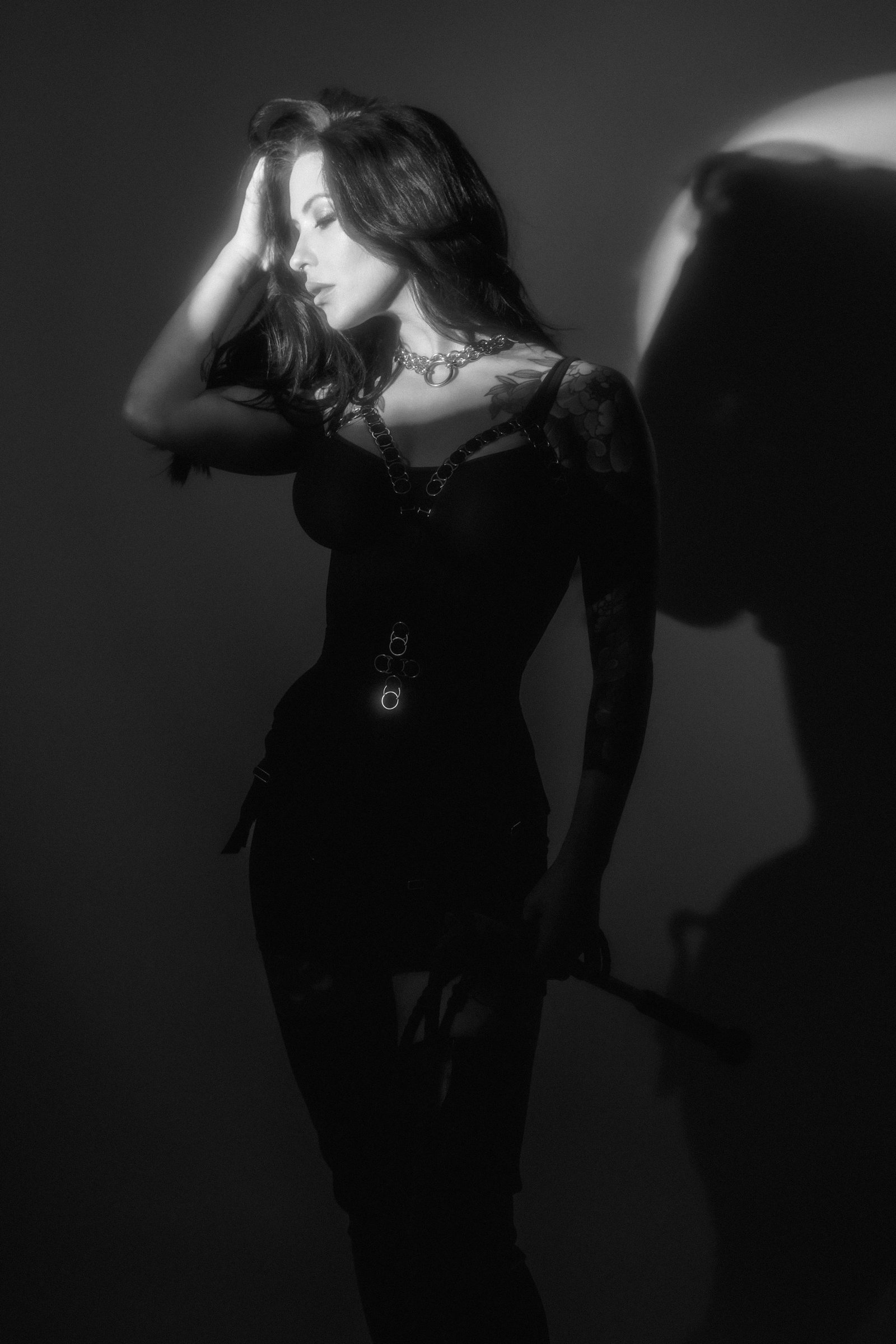Black and white photo of woman in dark outfit with tattoo on arm, holding a whip, in soft light.