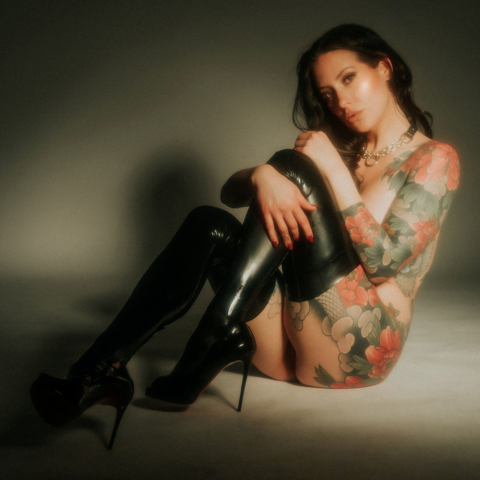 Tattooed woman with floral sleeve sit wearing shiny black thigh-high boots and a chain necklace.