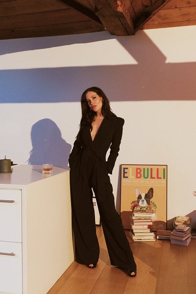 Woman in a black jumpsuit leaning against a white counter in a sunlit room with books and a poster.