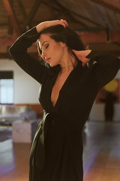 Woman in a black dress with eyes closed, posing with hands behind her head indoors.